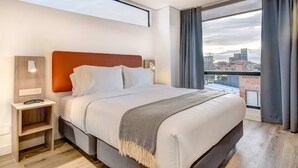 A modern hotel room with a large bed, a nightstand, and a city view through the window. - w* | Radiant 2BR w/ Balcony in Santa Barbara (Bogotá)
