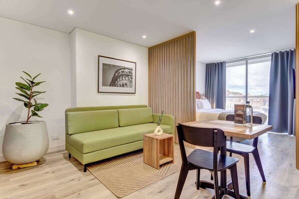 Un salon moderne avec un canapé vert, une table basse en bois et un coin repas avec des chaises. On y trouve une grande plante en pot, un tableau encadré au mur et une vue sur la ville à travers de grandes fenêtres.