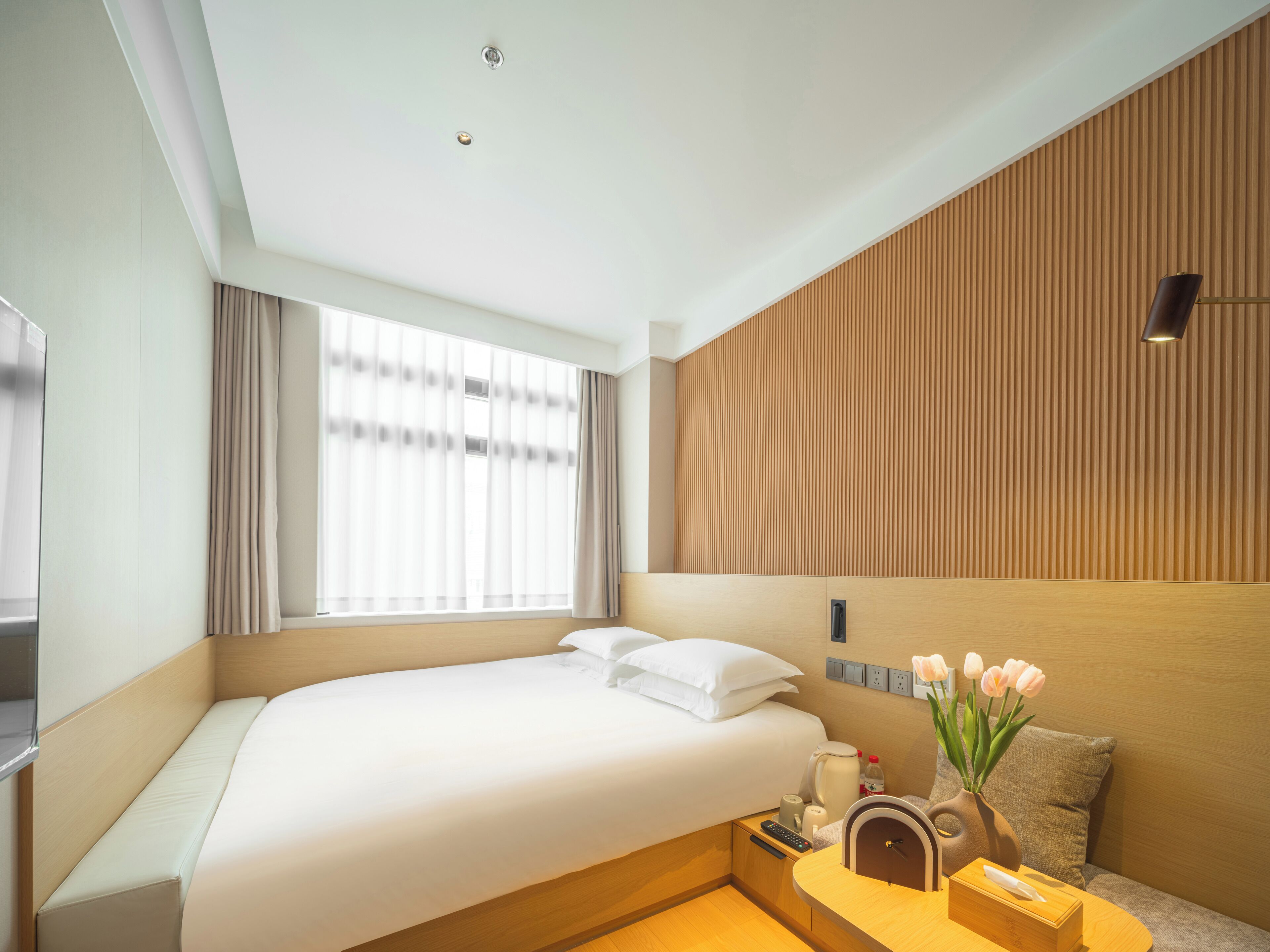 A modern hotel room with a large bed, a bedside table with a vase of flowers, a wooden headboard, and a large window with curtains.
