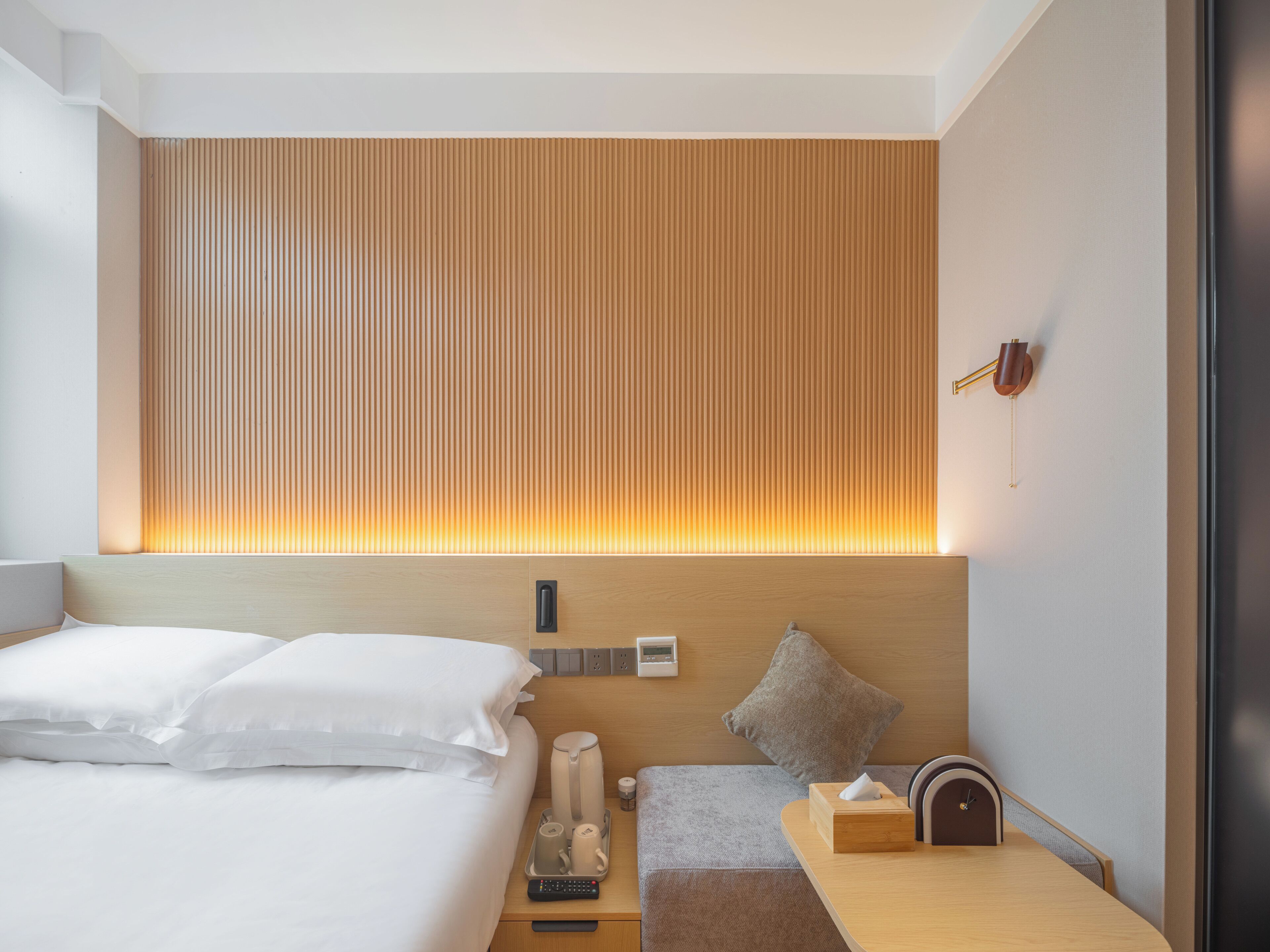 Una habitación de hotel moderna con una cama bien tendida, una mesita de noche con lámpara, una pequeña mesa con un reloj y un panel de control en la pared.