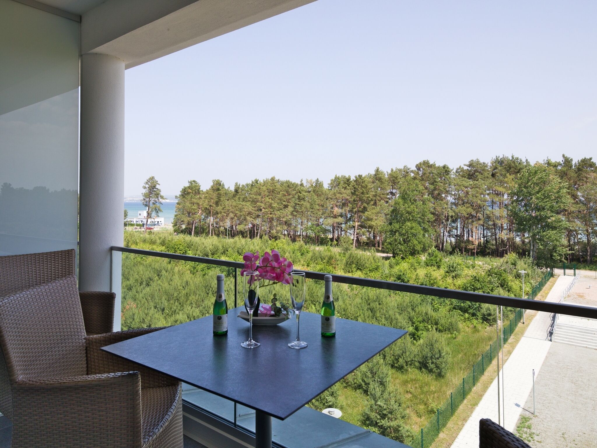 A balcony with wicker furniture, a table with two wine glasses and bottles, and a view of a forest and beach.