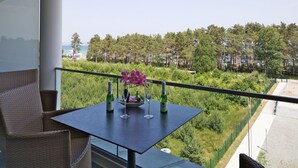 A balcony with wicker furniture, a table with two wine glasses and bottles, and a view of a forest and beach.