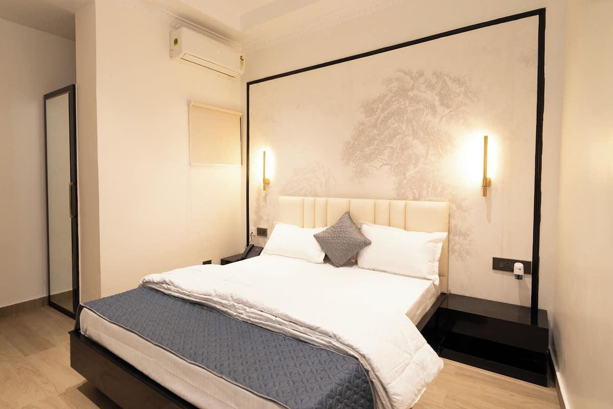 A modern hotel room with a large bed, a bedside table, a chair, and a wall-mounted air conditioning unit.