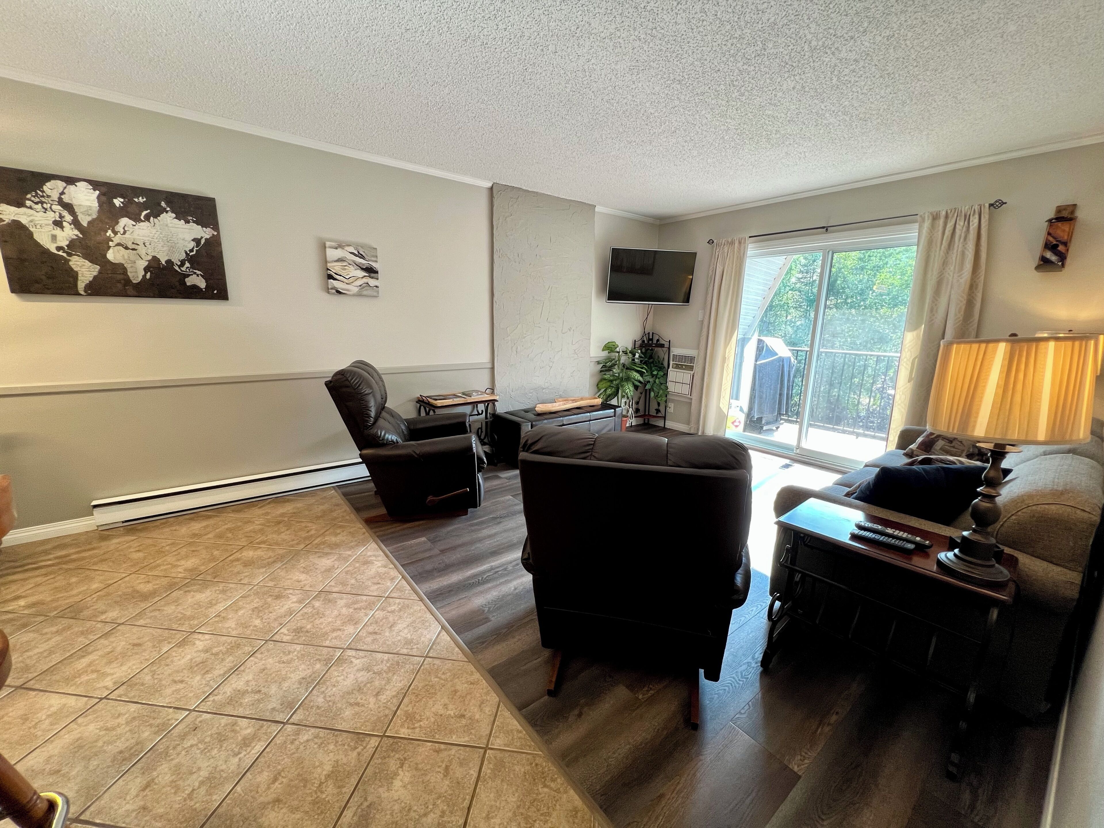 A living room with a black leather sofa, a black recliner, a wooden coffee table, a TV mounted on the wall, and a large window with curtains.