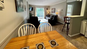 A dining area with a wooden table and chairs, a kitchen area with a bar stool, and a living room with a sofa and a TV. - Ski, Bike, Golf, Repeat (Kimberley)
