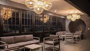 A modern lounge area with a sofa, chairs, and tables, large windows with city views, and decorative hanging lights. - Cozy Apartment in San José Costa Rica With Stunning Sunset Views (San José)