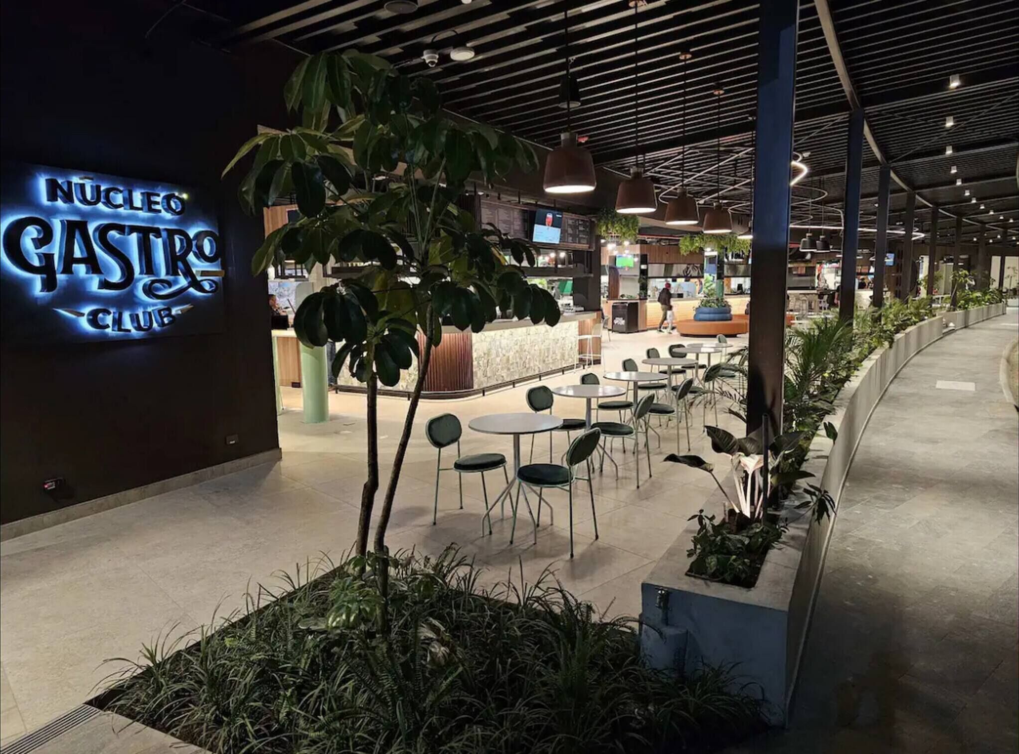 An indoor dining area with tables and chairs, a neon sign for 'Nucleo Gastros Club', and indoor plants.