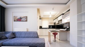 Una sala moderna con un sofá gris, una mesita blanca y un taburete rojo. Se ve una cocina al fondo, con armarios blancos y una encimera negra.