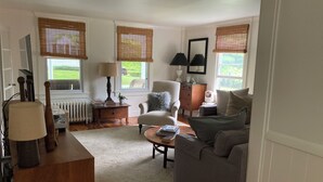 Una sala de estar con sofá, sillón, mesa de centro y lámpara. Se ve una ventana con persianas, un radiador y un espejo en la pared.