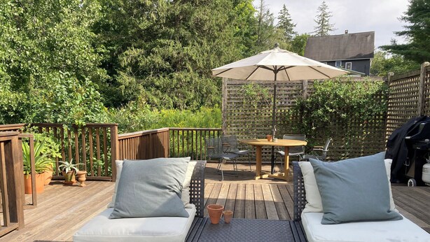Una terraza de madera con mobiliario exterior, incluyendo dos sillones con cojines y una mesa, rodeada de follaje y una cerca de madera.