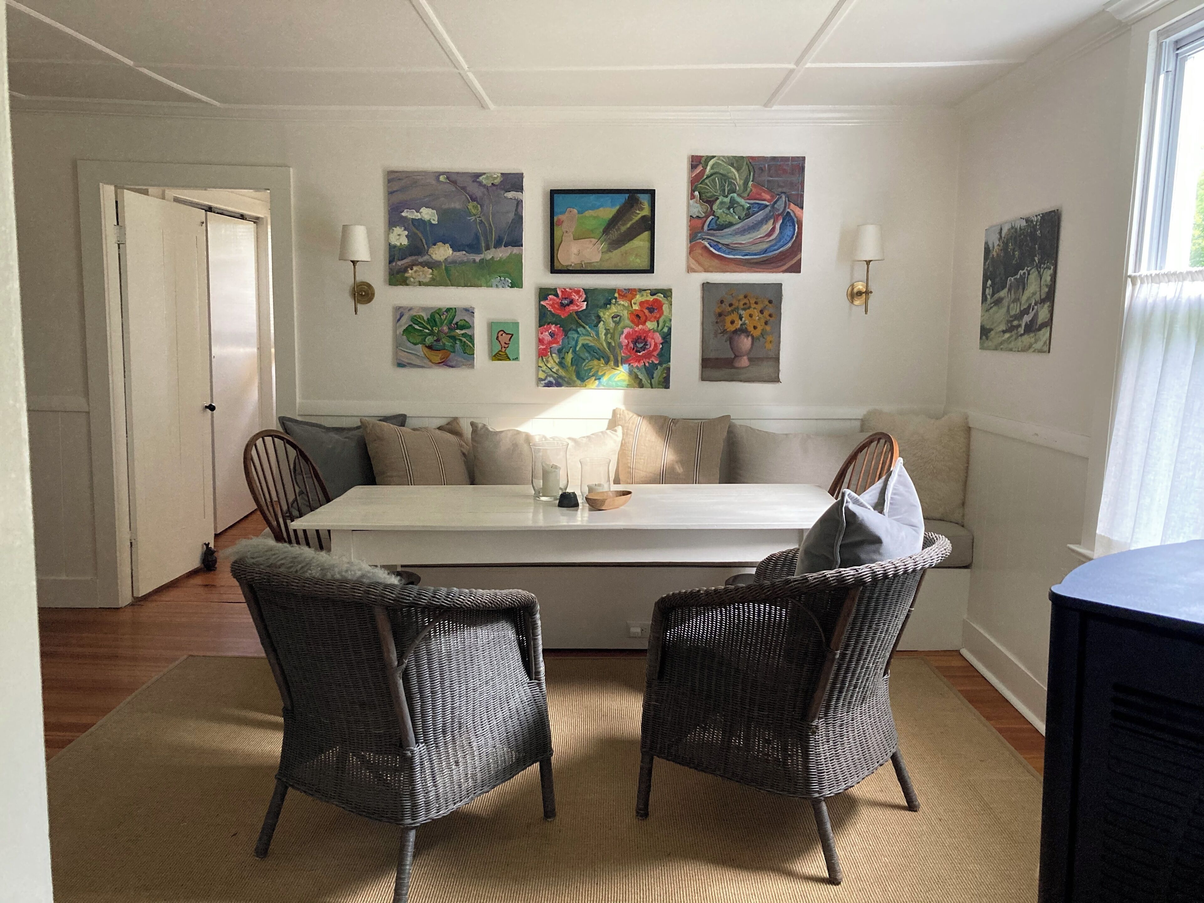A dining area with a white table, two wicker chairs, and a bench. There are several paintings on the wall, and a door is visible to the left.