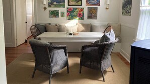 Un comedor con una mesa blanca, dos sillas de mimbre y un banco. Hay varias pinturas en la pared, y se ve una puerta a la izquierda.