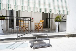 Uma varanda com duas cadeiras de madeira, uma mesa pequena e um banco de metal.