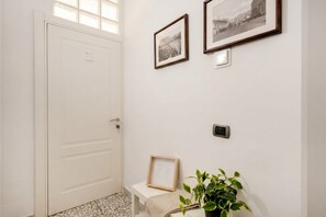 Um corredor com uma porta branca, um banco, duas imagens emolduradas na parede e um vaso de planta.