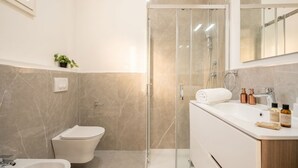 A modern bathroom with a glass shower enclosure, a white toilet, a sink with toiletries, and a mirror above the sink.