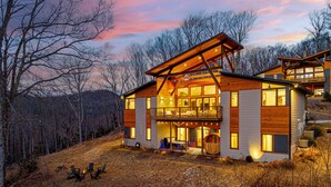 A modern two-story house with a large deck and a fireplace, surrounded by a wooded area.