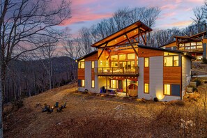 A modern two-story house with a large deck and a fireplace, surrounded by a wooded area.