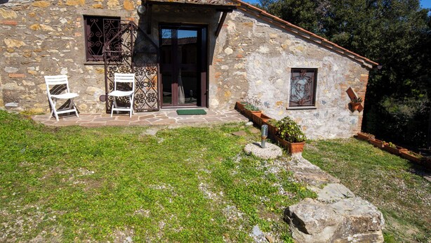 Un edificio de piedra con techo de tejas, una puerta de madera y una ventana pequeña. Hay dos sillas blancas afuera y una planta en maceta cerca de la entrada.