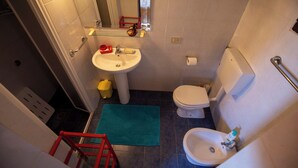 Un baño pequeño con lavamanos, inodoro y bidé. Hay una escalera roja y un tapete verde en el suelo.