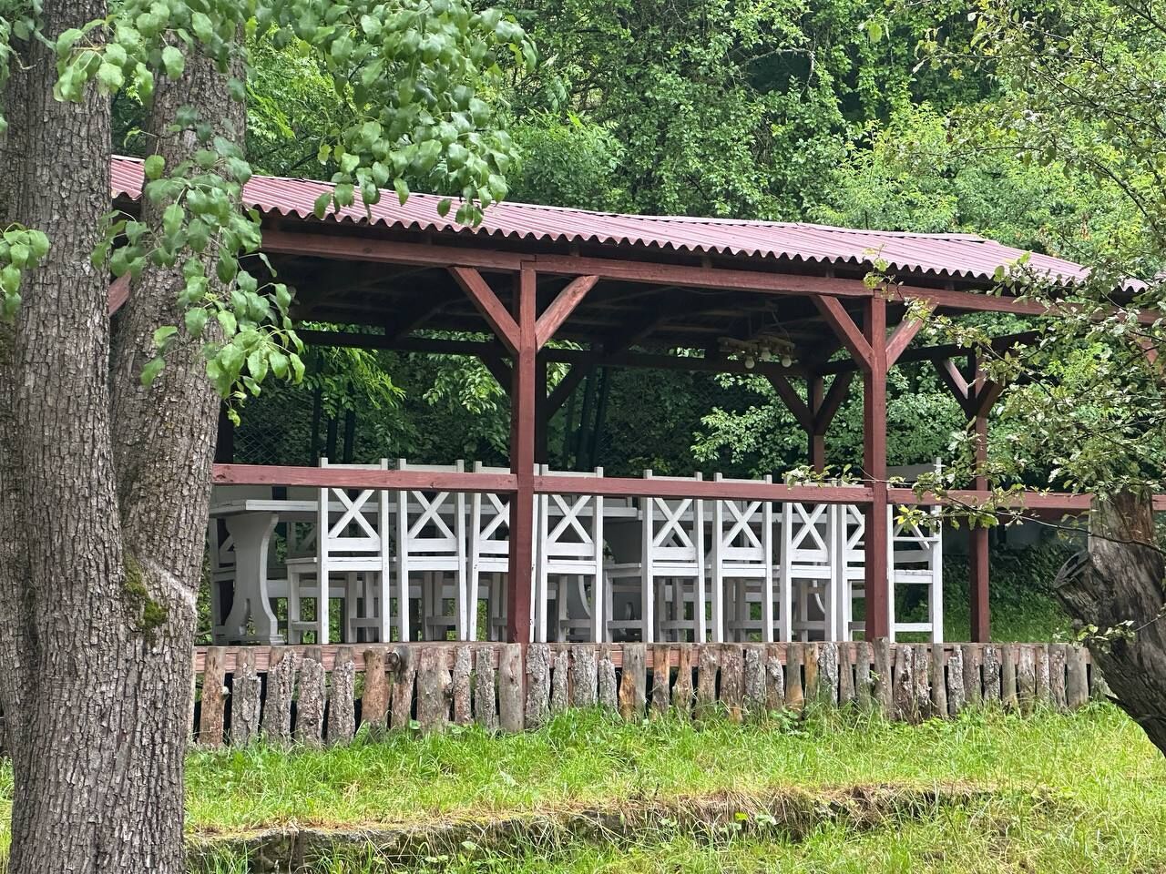 Un pabellón de madera con techo rojo, rodeado de una cerca de celosía blanca, en un entorno verde exuberante.