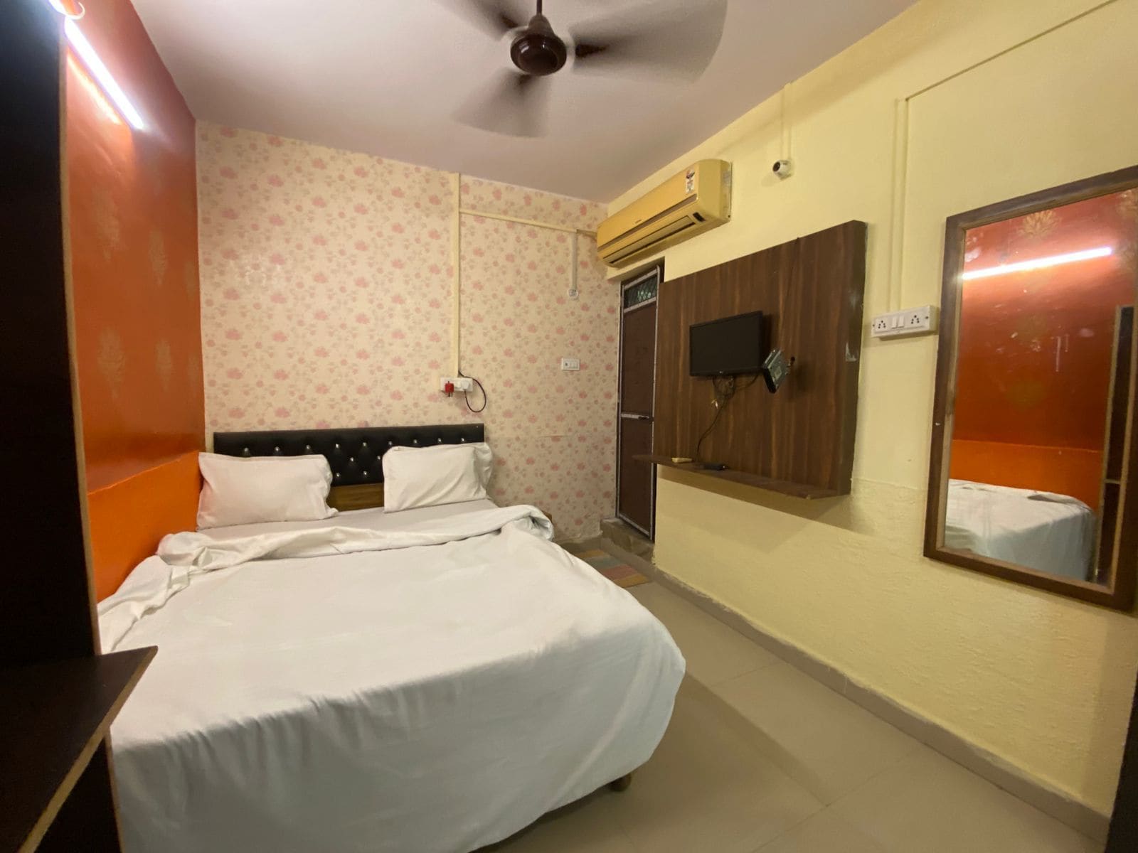 Een hotelkamer met een bed, een aan de muur bevestigde televisie, een airconditioning en een spiegel die een deel van de kamer weerspiegelt.