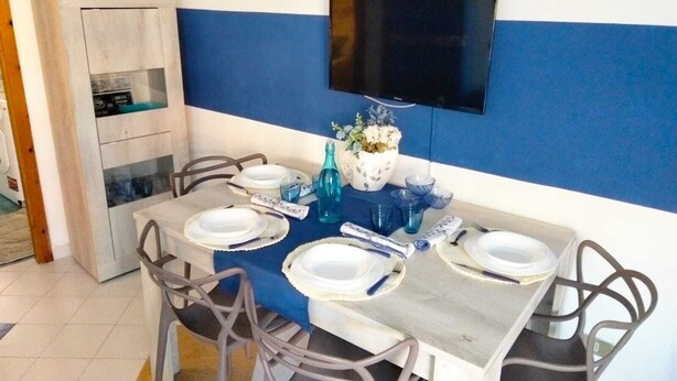 A dining area with a blue and white striped wall, a flat-screen TV mounted above the table, and a cabinet with glass doors to the left. - BLUE MARINE: cozy apartment 200 meters from the sea (Ugento)