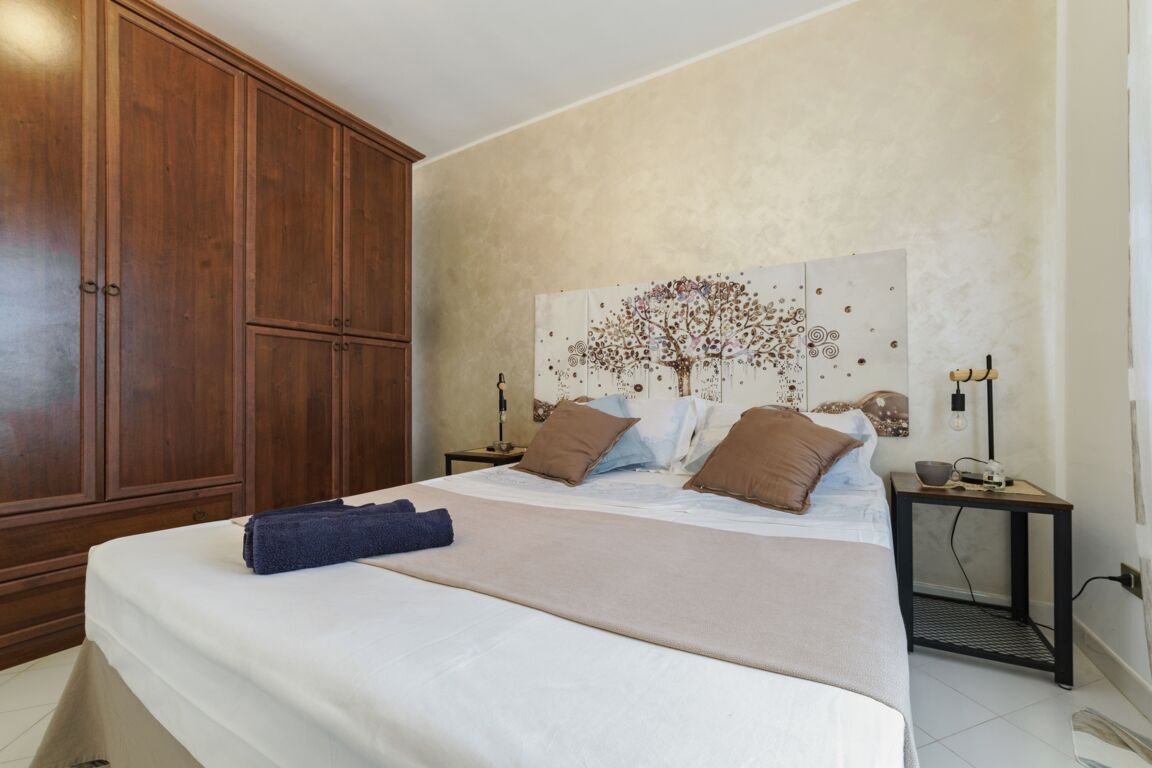 Ein Schlafzimmer mit großem Holzschrank, einem Bett mit beiger Bettdecke und mehreren Kissen, einem Wandbild und einem kleinen Nachttisch mit Lampe.