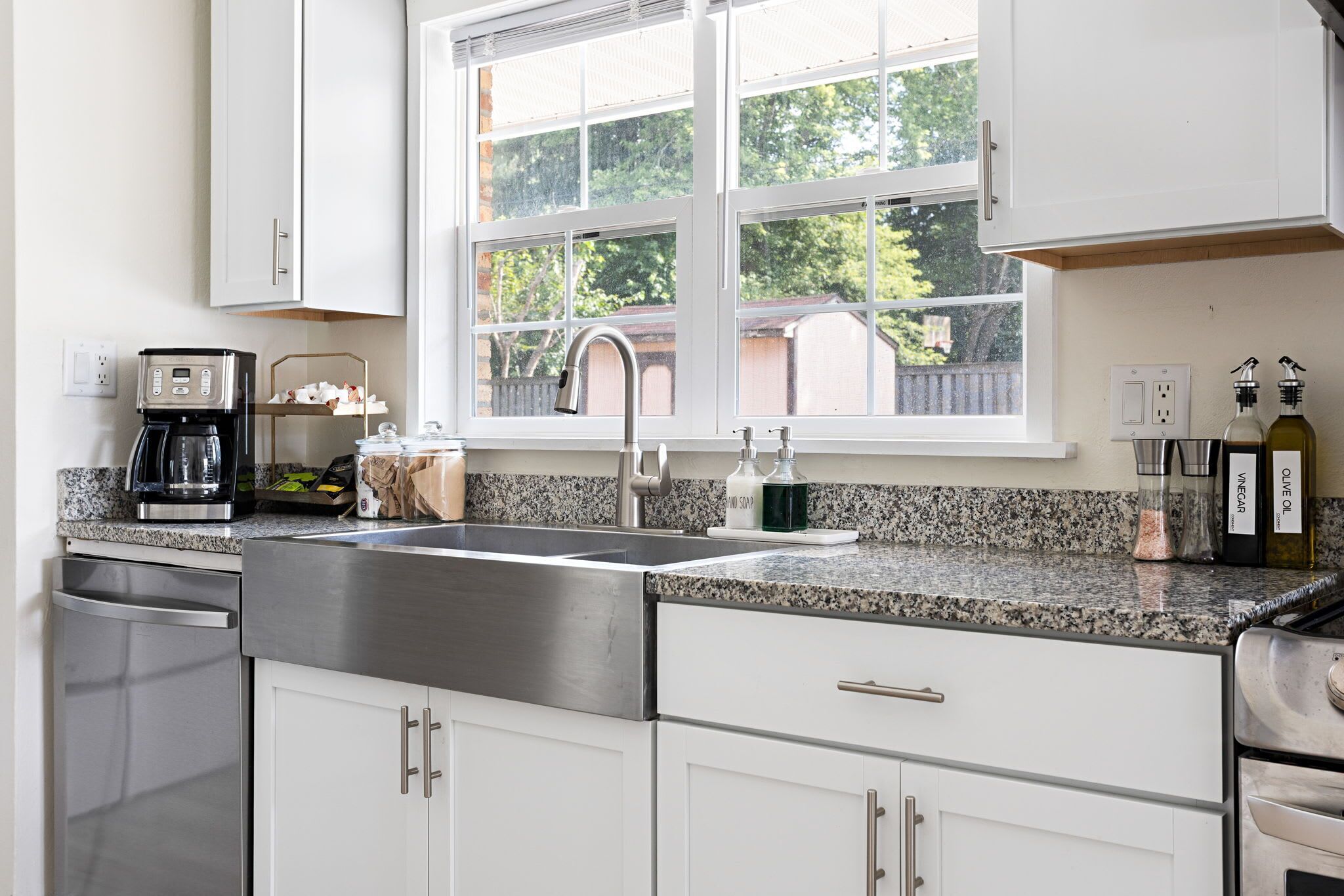 A modern kitchen with white cabinets, a stainless steel sink, a coffee maker, and a window with a view of trees.