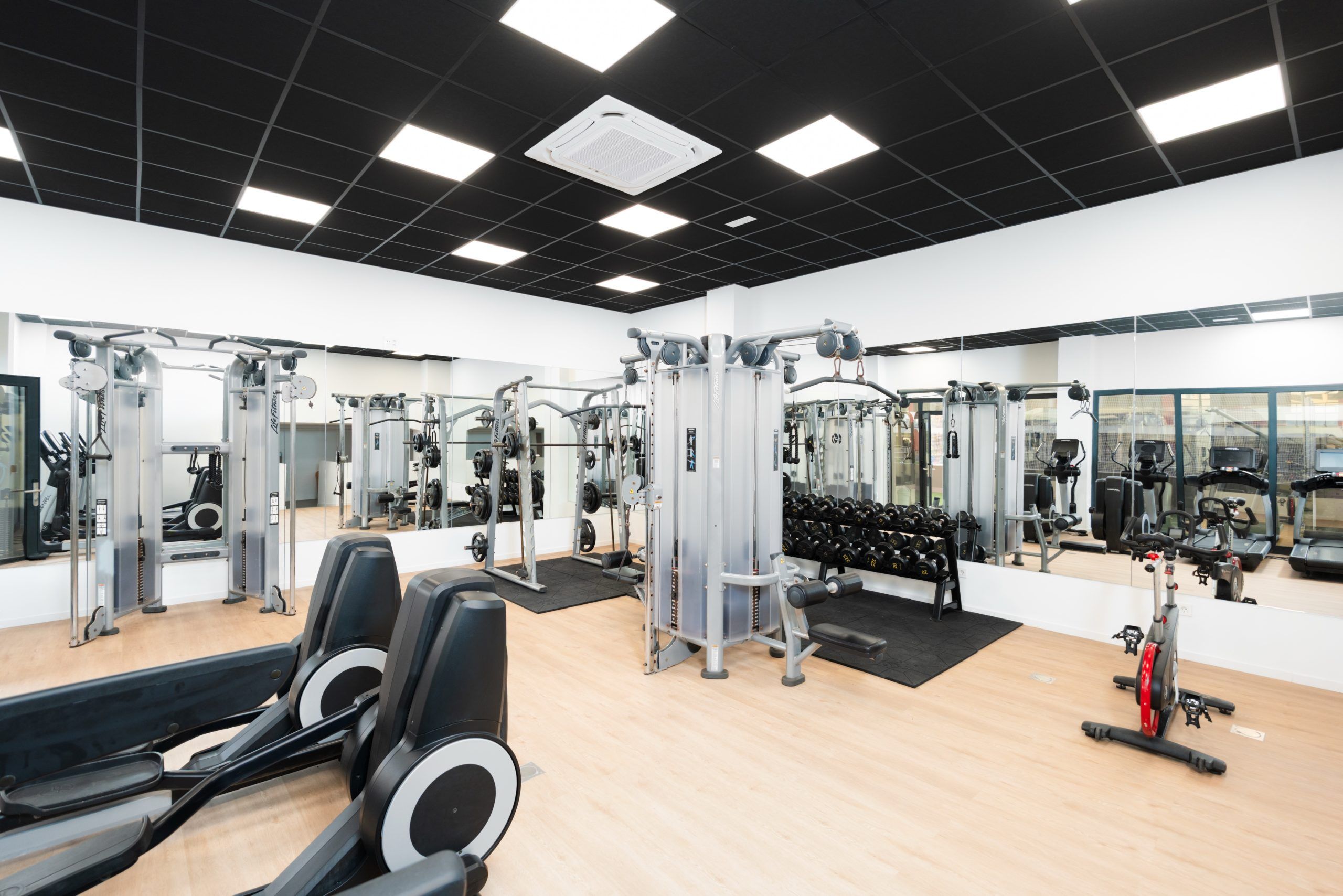 A gym with various exercise equipment, including weight machines and free weights, and a mirror wall.