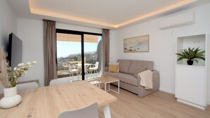 A modern living room with a sofa, a dining table, and a TV. There is a balcony with a view of the city. - Natural Park (Mogán)