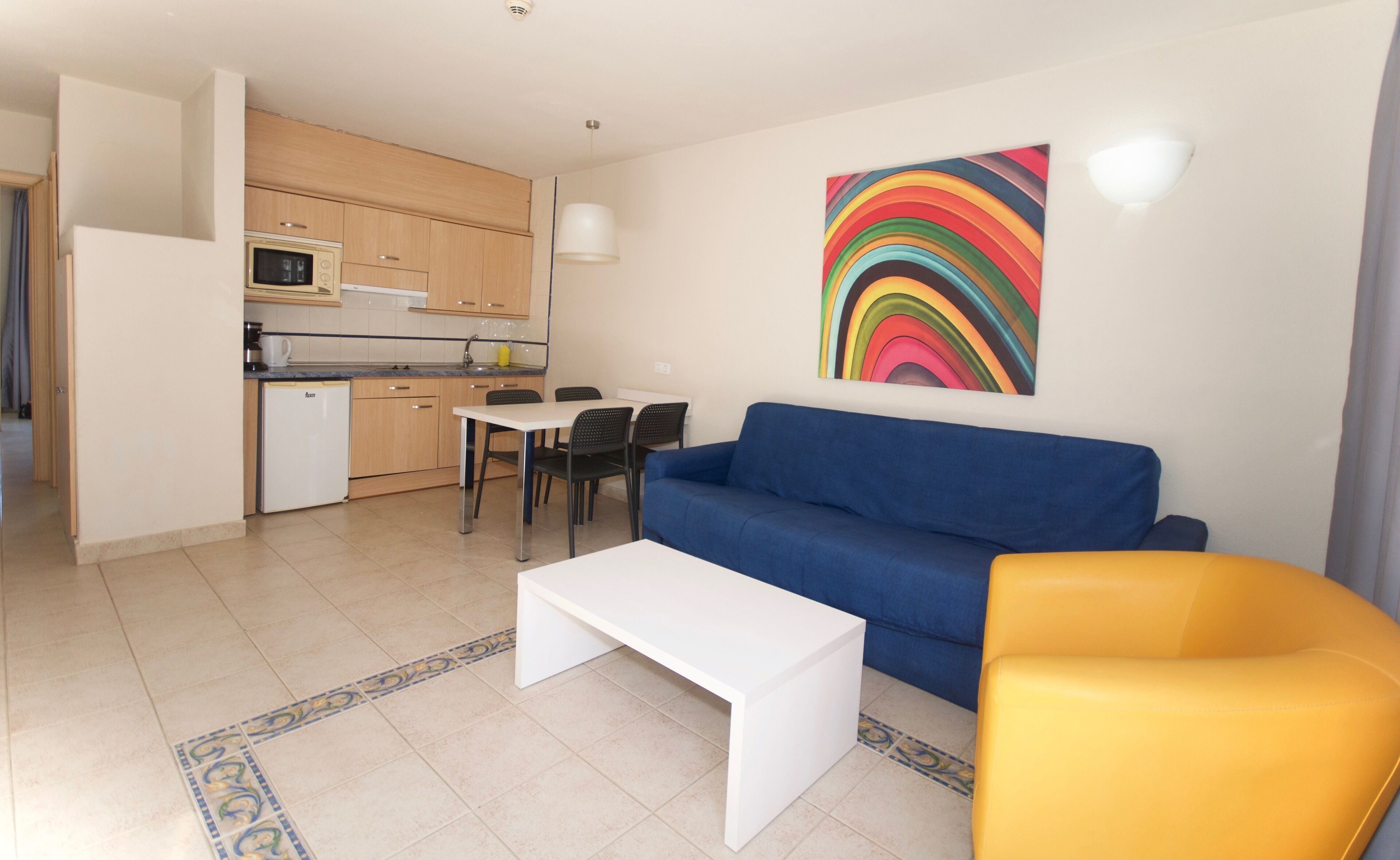 A compact living space with a kitchenette, a blue sofa, a white coffee table, and a yellow armchair.