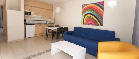 A compact living space with a kitchenette, a blue sofa, a white coffee table, and a yellow armchair.
