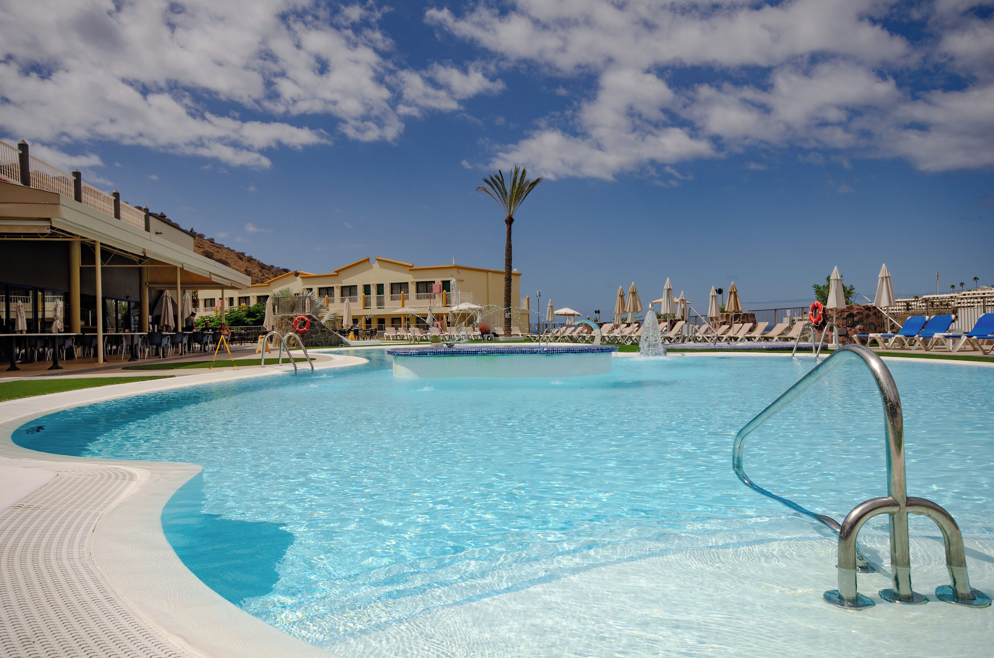 A large outdoor swimming pool with a palm tree, lounge chairs, and a building with a balcony in the background.