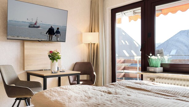 A hotel room with a bed, a desk with a vase of flowers, a chair, a television mounted on the wall, and a window with a view of buildings. - Quartier 16<br>Apartment in a quiet location and access to sea time (Büsum)