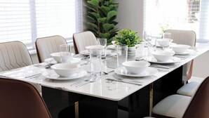 Mesa de comedor lista para seis personas, con platos blancos, boles y vasos transparentes, rodeada de sillas marrones.