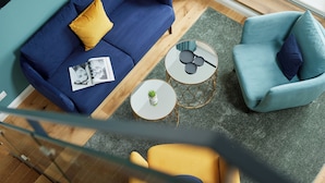A modern living room with a blue sofa, a yellow armchair, and a teal armchair. There are two round tables, one with a small plant and the other with a decorative item. - Completion 2025: Exclusive accommodation with balcony close to the beach! (Sankt Peter-Ording)