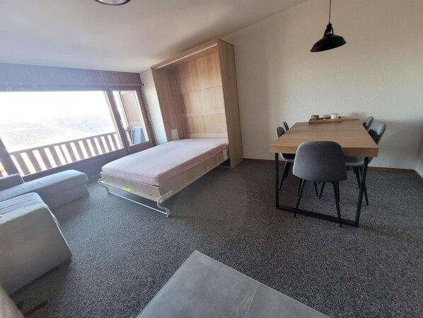 A compact room with a bed, a dining table with chairs, and a view of the outdoors.