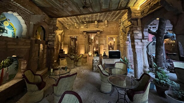 A rustic interior with stone walls, wooden beams, and traditional decor. There are wicker chairs and a small table, with a decorative elephant sculpture on the left. - Jhankar Haveli (Jodhpur)