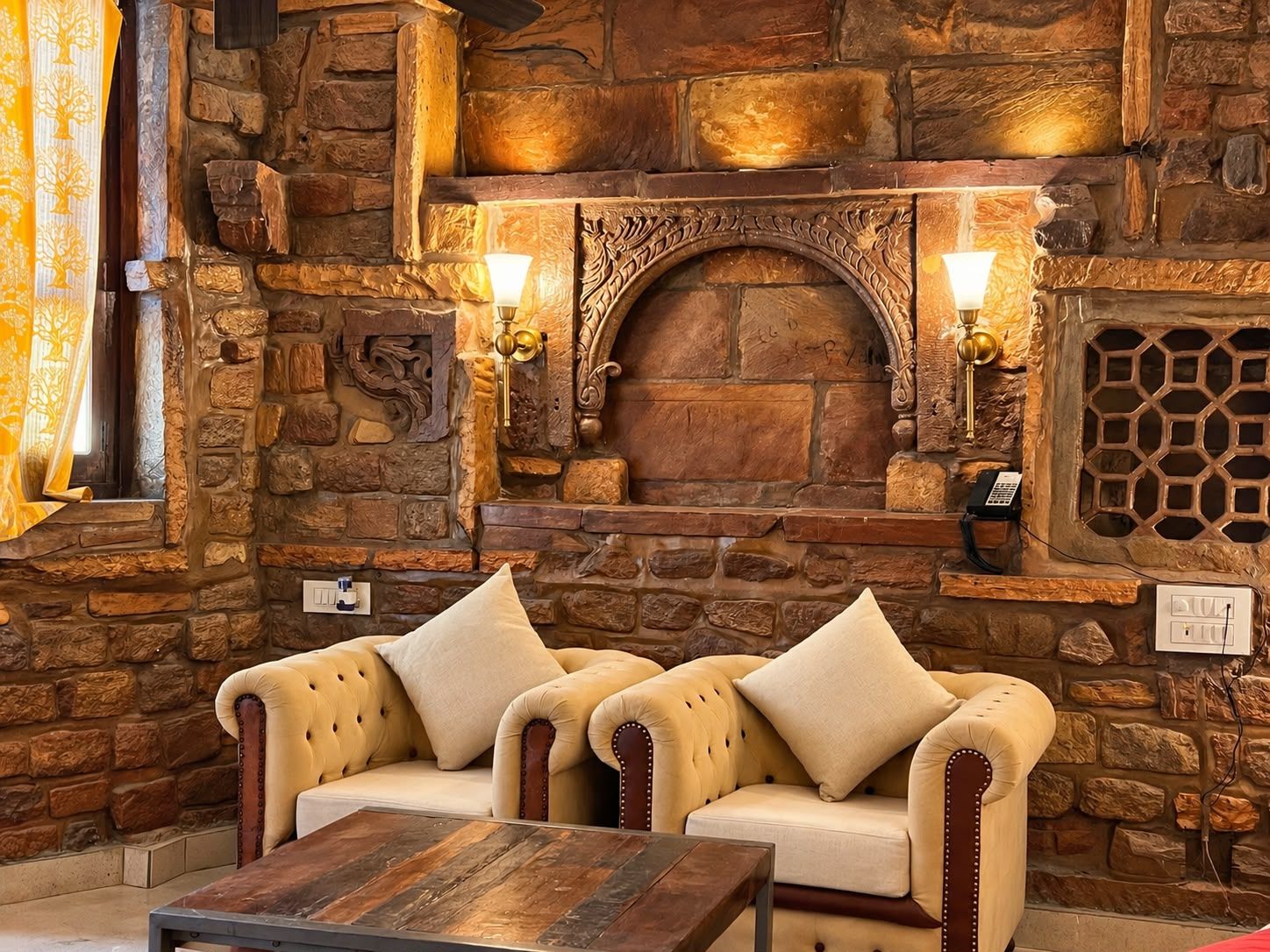 Una chimenea de piedra con dos lámparas de pared, un sofá con cojines y una mesa de centro de madera, además de un panel decorativo en la pared.