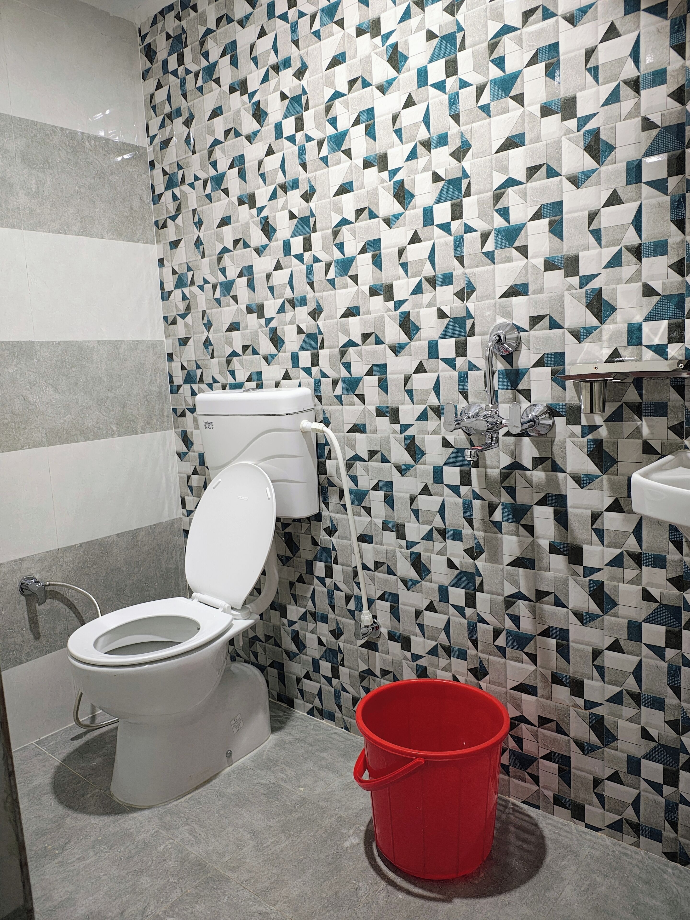 A bathroom with a toilet, a red bucket, and a shower with a geometric tile pattern.