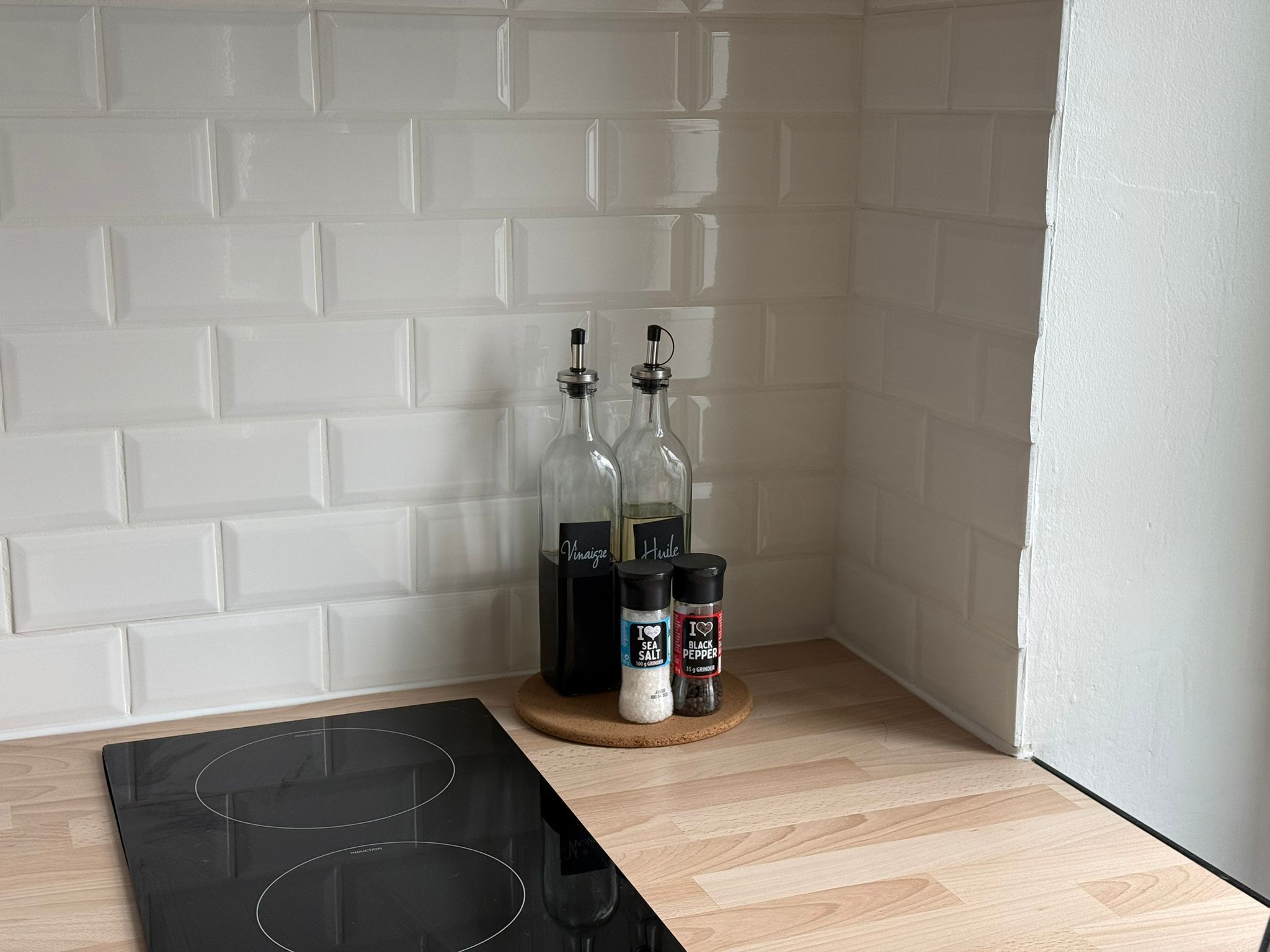 Una cocina con baldosas blancas tipo metro, una placa de inducción negra y dos botellas de vino sobre una tabla de madera.