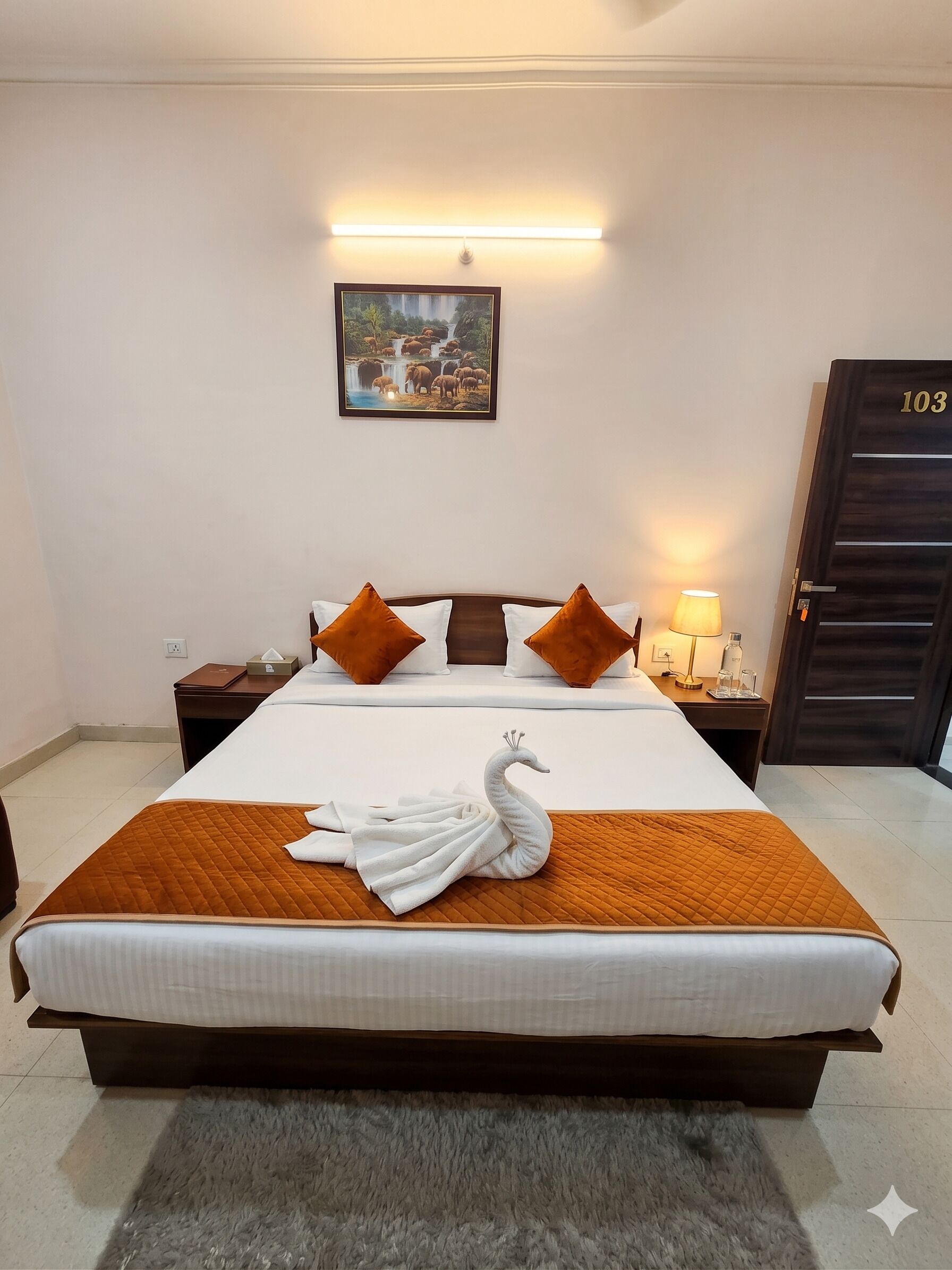 A hotel room with a bed, a nightstand, a lamp, a framed picture on the wall, and a door with a number.