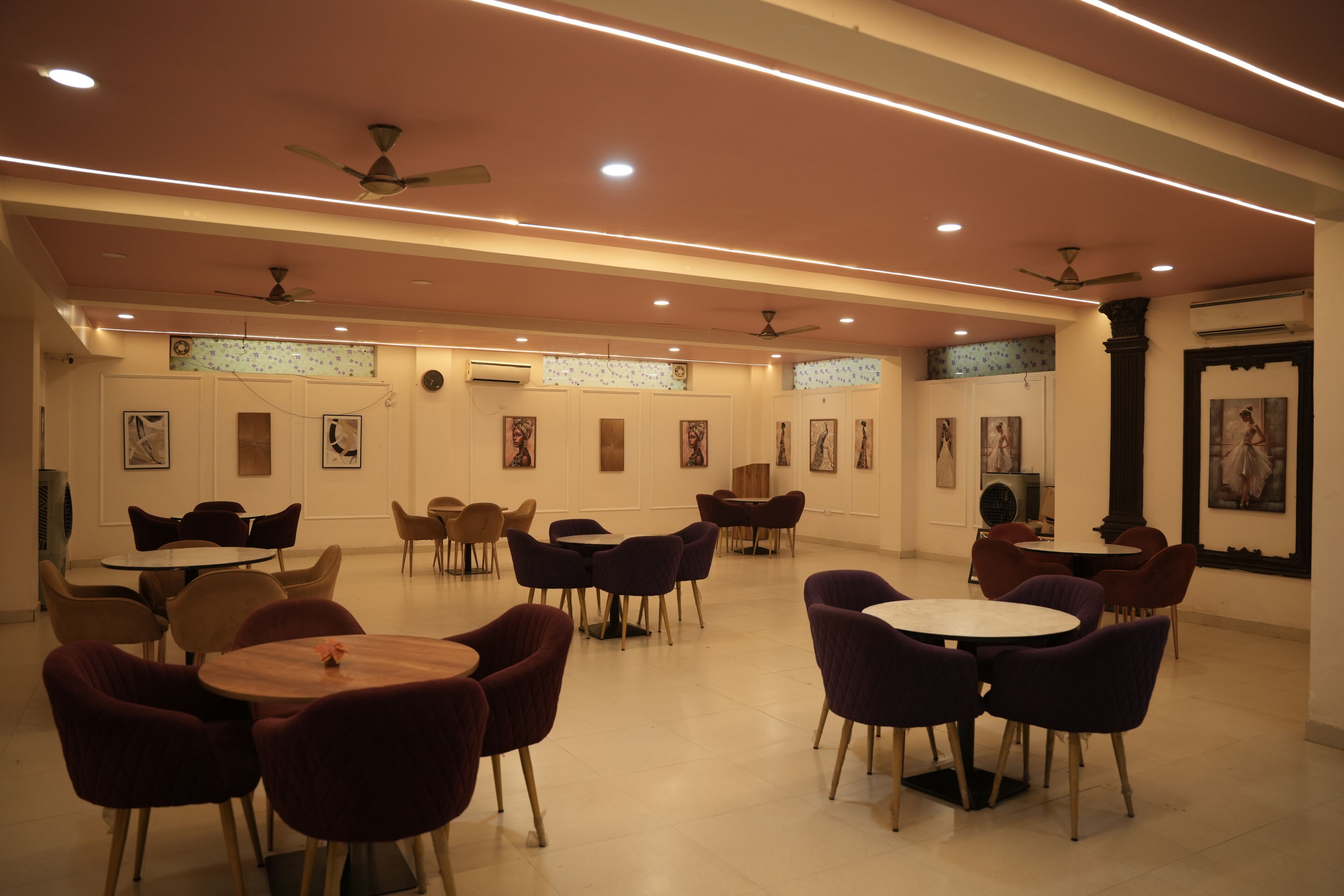 A spacious dining area with multiple round tables and chairs, ceiling fans, and wall-mounted air conditioning units.