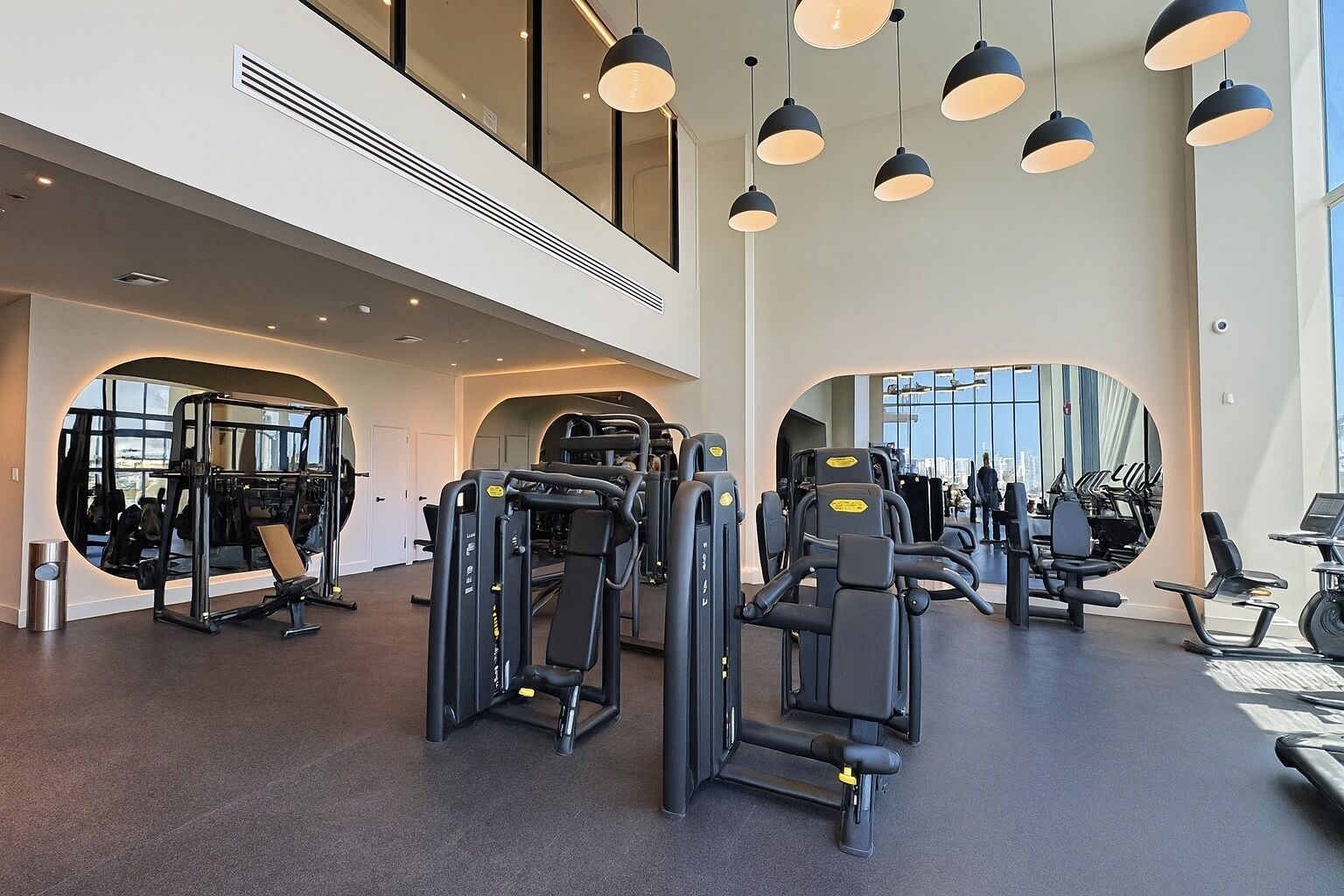 Uma academia moderna com diversos equipamentos de exercício, amplas janelas e iluminação pendente.