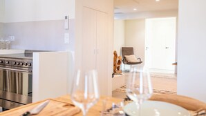 A modern kitchen with stainless steel appliances, a dining area with a wooden table and wine glasses, and a view into a living room with a sofa and a door.