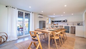 A modern kitchen with a dining area, featuring a large table, wooden chairs, and a view through glass doors.