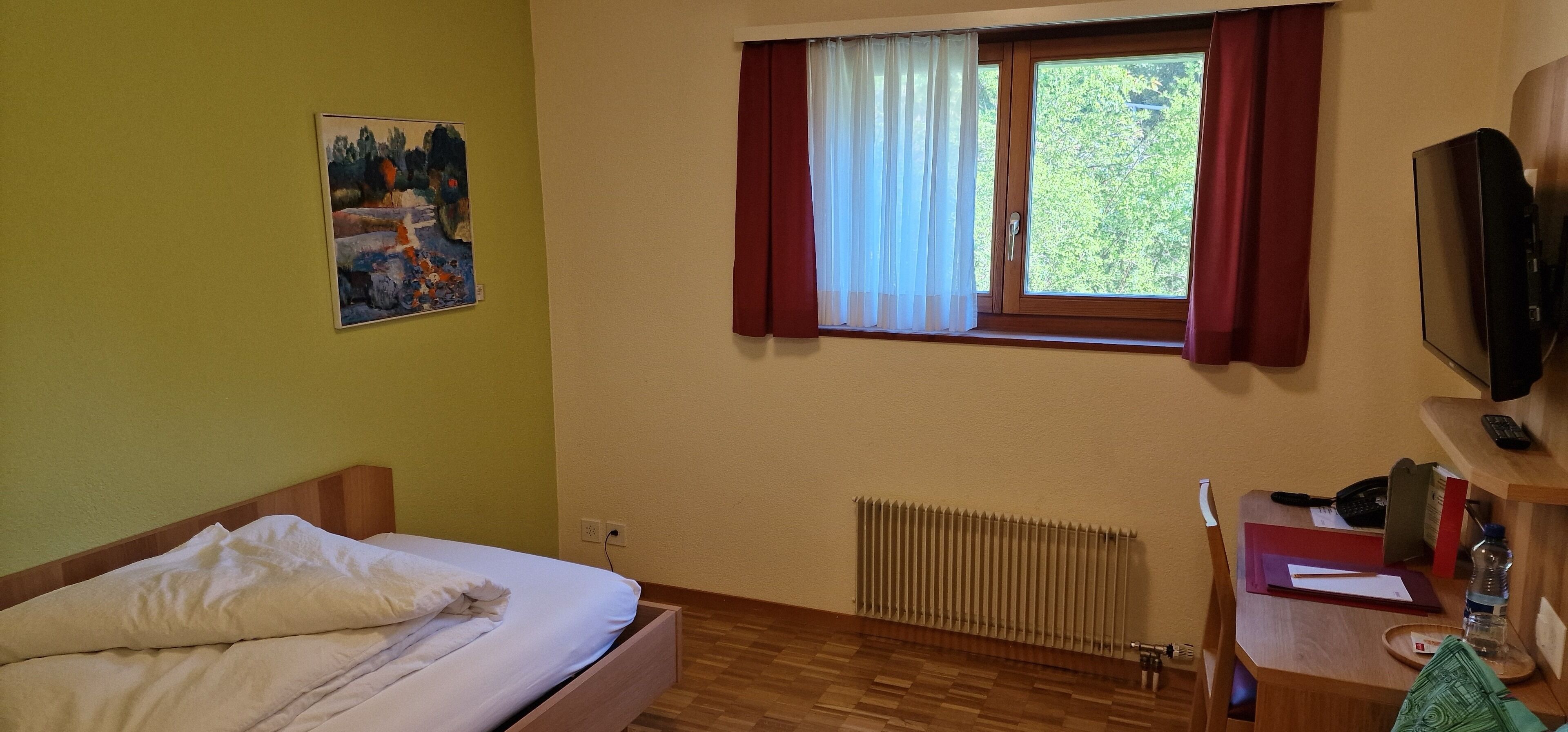 Une chambre à coucher avec un lit, une fenêtre avec des rideaux, un radiateur, une télévision et un bureau avec un téléphone et un livre.