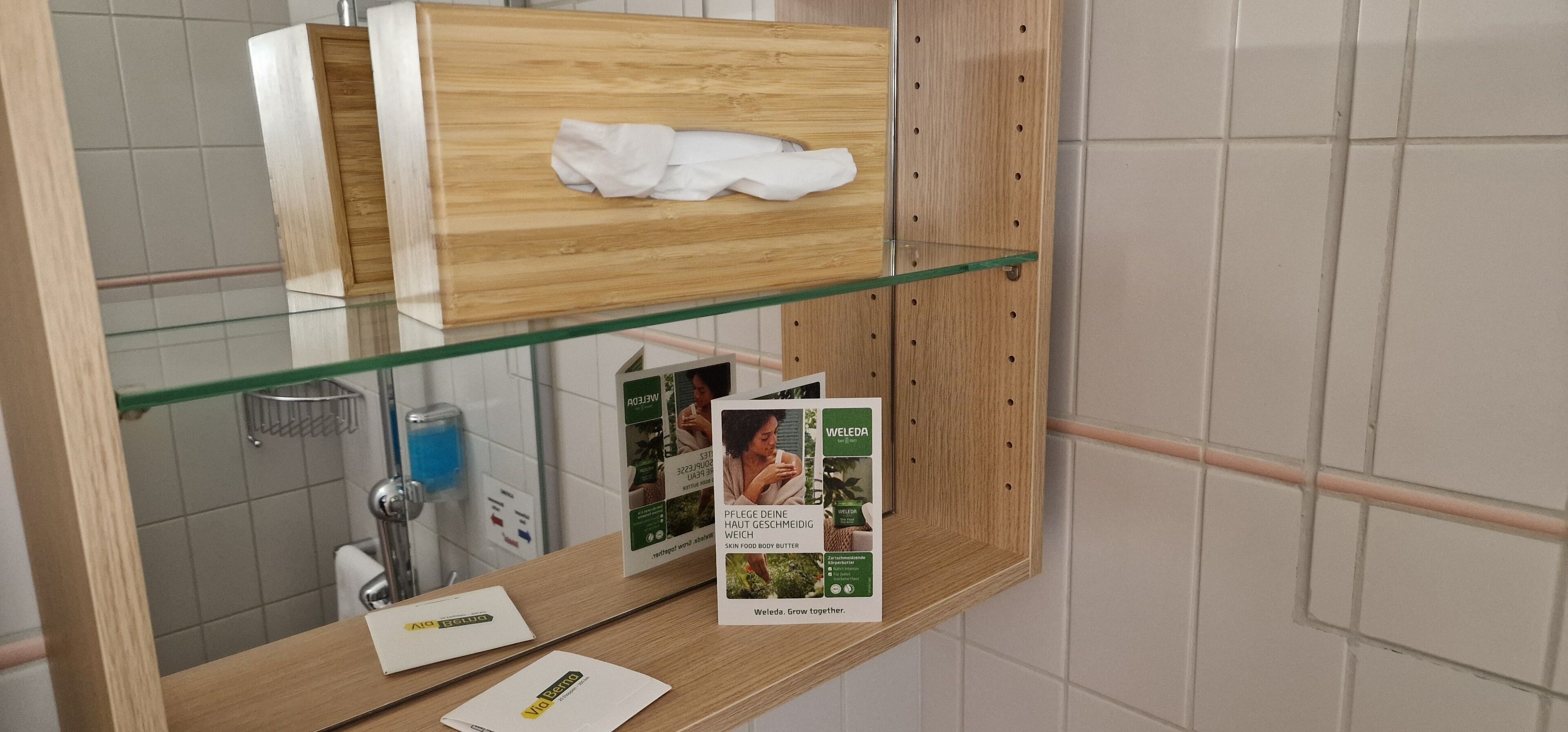 A bathroom with a glass shelf holding a white towel and a tissue box, a glass shower enclosure, and a toilet paper roll.