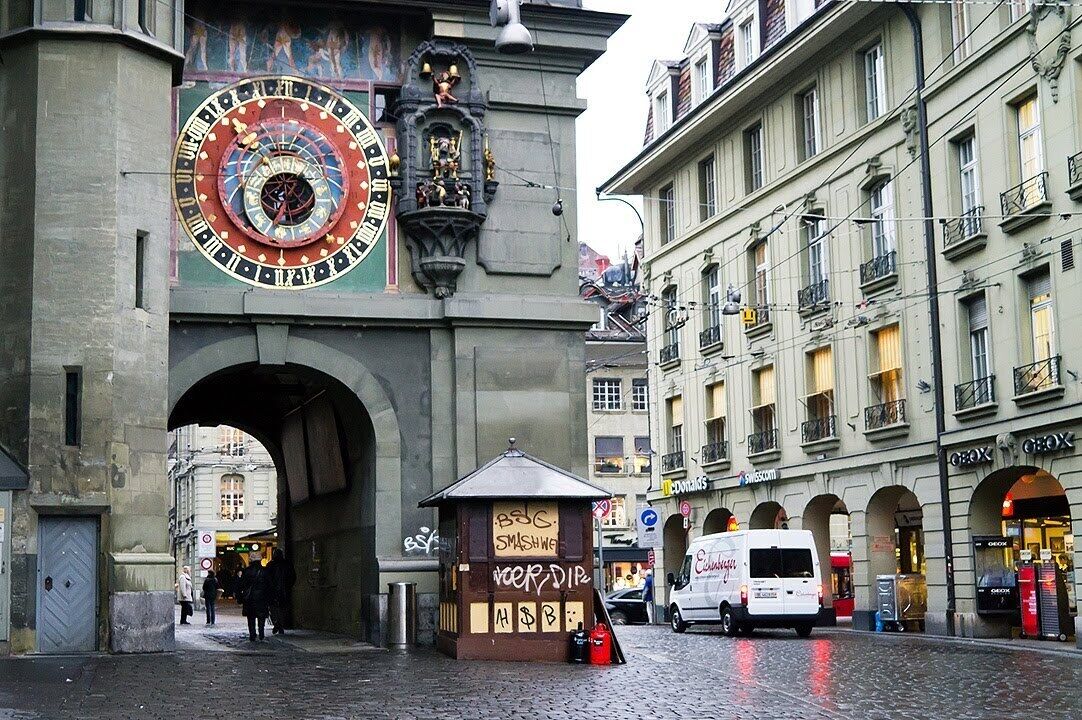 An urban street scene with a large, ornate clock tower, a small kiosk, and storefronts.