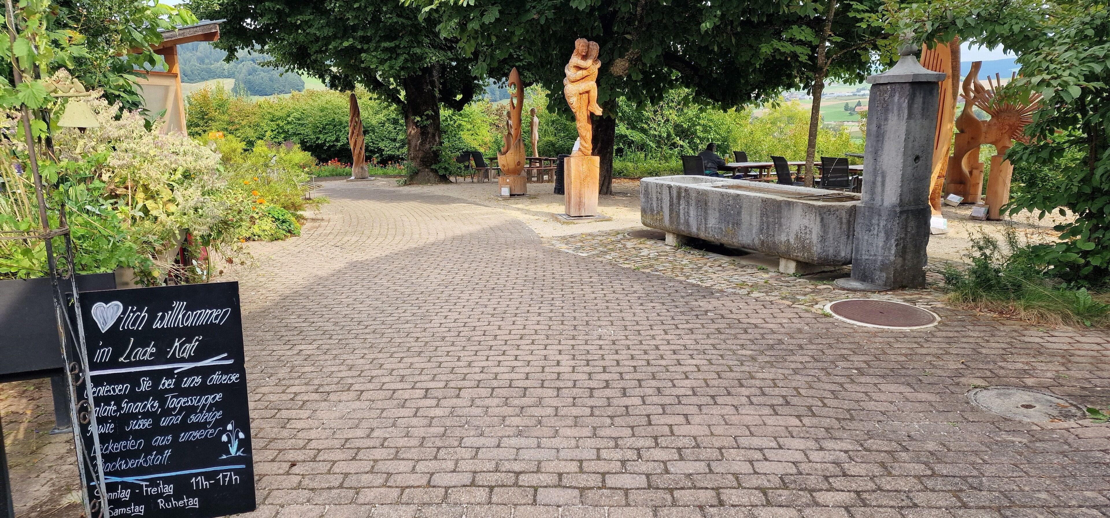 Un sendero pavimentado que lleva a una zona de descanso con esculturas artísticas y un cartel.